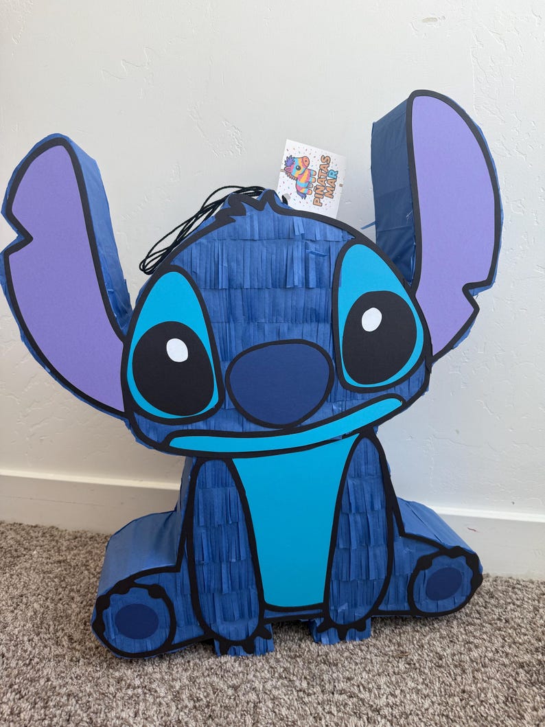 May include: A large, blue Stitch pi&ntilde;ata with purple ears and a light blue belly. The pi&ntilde;ata is made of crepe paper and has black eyes and a black nose. A tag with the words "Pi&ntilde;atas Mar" is attached.