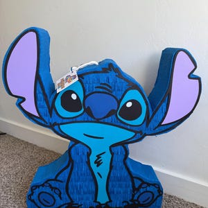 May include: A blue pi&ntilde;ata in the shape of the character Stitch, with large ears, big eyes, and a wide smile. The pi&ntilde;ata is made of blue crepe paper with black outlines and purple accents on the ears. A tag hangs from the top.