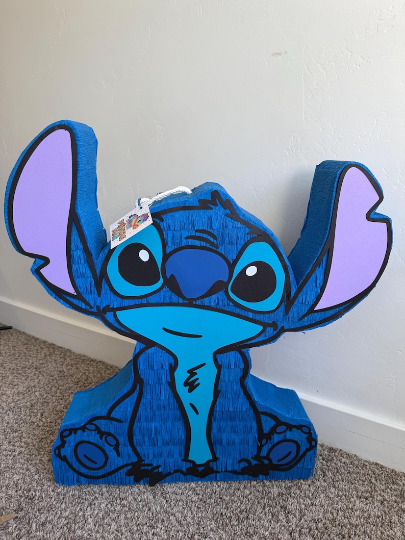 May include: A blue pi&ntilde;ata in the shape of the character Stitch, with large ears, big eyes, and a wide smile. The pi&ntilde;ata is made of blue crepe paper with black outlines and purple accents on the ears. A tag hangs from the top.