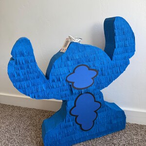 May include: A large, blue pi&ntilde;ata shaped like a cartoon character. It's made from blue crepe paper, featuring two blue cloud shapes outlined in black. The pi&ntilde;ata has large ears and a tag attached to the top.
