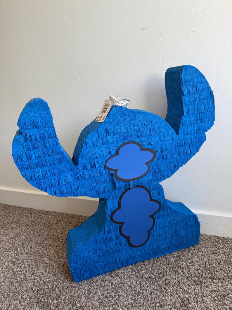 May include: A large, blue pi&ntilde;ata shaped like a cartoon character. It's made from blue crepe paper, featuring two blue cloud shapes outlined in black. The pi&ntilde;ata has large ears and a tag attached to the top.