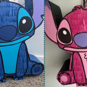 May include: Two pi&ntilde;atas shaped like the cartoon character Stitch. One is blue with light blue details, the other is pink with black accents. Both have large ears, big eyes, and a friendly expression. They are made of paper and are likely for a party or celebration.
