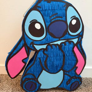 May include: A large, blue pi&ntilde;ata shaped like a cartoon character with big eyes, pink ears, and a light blue belly. The pi&ntilde;ata is made of blue crepe paper and has a rope for hanging. The character's face is detailed with black outlines.