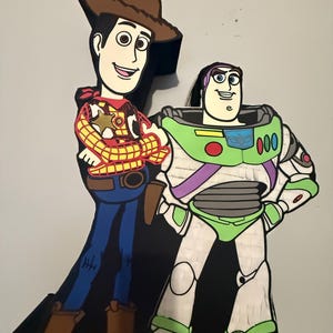 May include: A colorful pi&ntilde;ata featuring the characters Woody and Buzz Lightyear from the animated film Toy Story. Woody is wearing a cowboy hat, vest, and boots. Buzz Lightyear is in a green and white spacesuit. The pi&ntilde;ata is against a light gray background.
