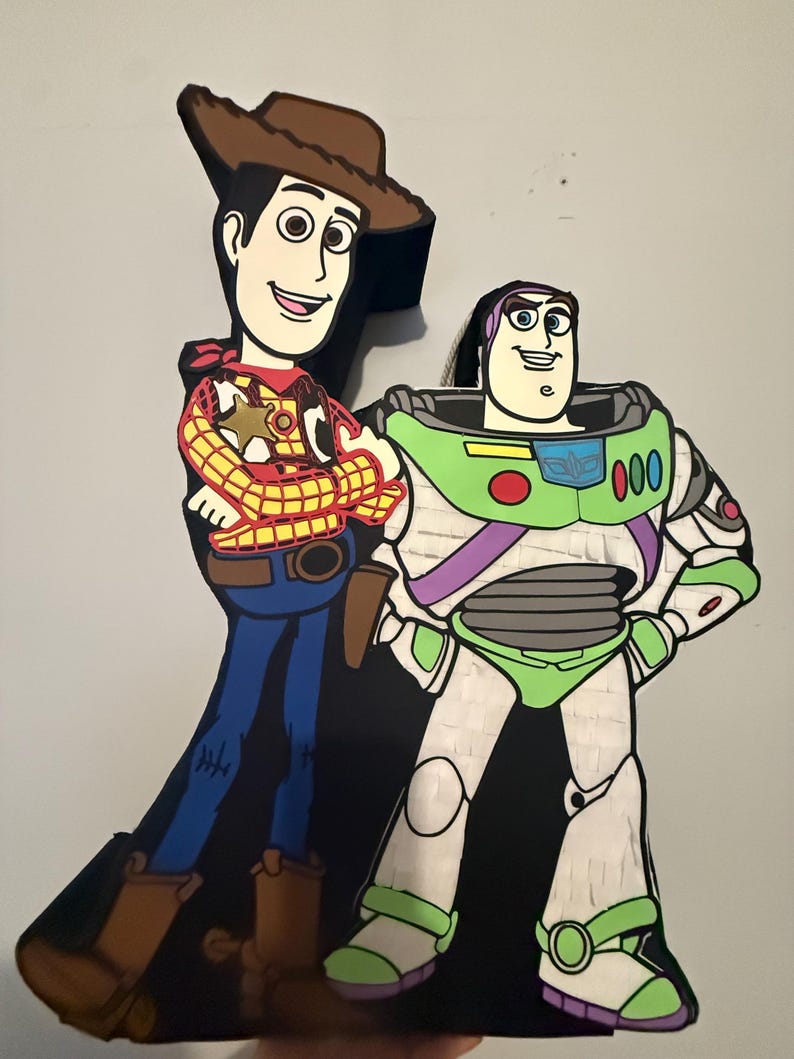 May include: A colorful pi&ntilde;ata featuring the characters Woody and Buzz Lightyear from the animated film Toy Story. Woody is wearing a cowboy hat, vest, and boots. Buzz Lightyear is in a green and white spacesuit. The pi&ntilde;ata is against a light gray background.