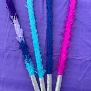 May include: Four colourful party decorations with fringed paper tops in shades of purple, blue, and pink. The fringed sections are attached to silver-coloured cylindrical handles. The decorations are arranged on a purple surface.