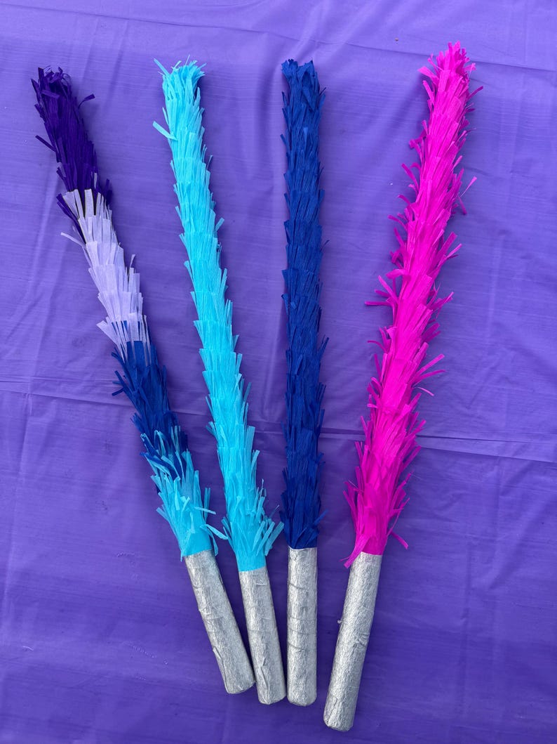 May include: Four colourful party decorations with fringed paper tops in shades of purple, blue, and pink. The fringed sections are attached to silver-coloured cylindrical handles. The decorations are arranged on a purple surface.