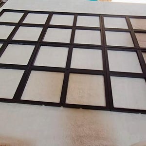 Könnte beinhalten: Eine schwarze gitterartige Struktur mit mehreren quadratischen Öffnungen, möglicherweise für ein Fenster oder eine Anzeige. Die Quadrate sind mit weißem und beigem Material gefüllt. Der Rahmen ist dunkel, und das Objekt liegt auf einer weißen Oberfläche.