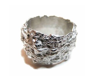 May include: A wide silver ring featuring a highly textured, irregular, and rugged surface that resembles organic, molten metal. The band has a thick, uneven profile with deep crevices and raised ridges throughout the entire exterior circumference.