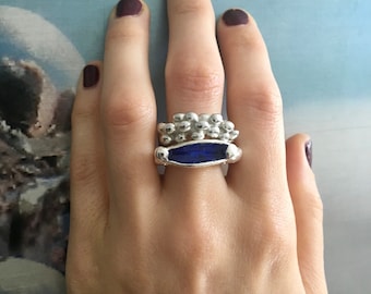May include: A silver ring set consisting of two stacked bands. The top band features a cluster of small, rounded silver spheres. The bottom band is a smooth silver setting holding a single, elongated, deep blue rectangular stone.