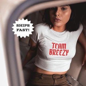 May include: White crop top with the text "Team Breezy" in red lettering. The model is wearing brown pants and is seated in a car. The image also includes a "Ships Fast!" graphic.