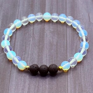 May include: A beaded bracelet featuring opalescent, light blue, and clear round beads. The bracelet also includes three dark, porous, round beads and small gold-colored beads. The bracelet is strung on a clear elastic cord.