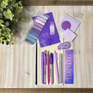 May include: A collection of purple stationery items, including pens, sticky notes, and a bookmark, arranged on a wooden surface. The items are in various shades of purple, with a few pops of pink and green. A small plant is in the upper left corner.