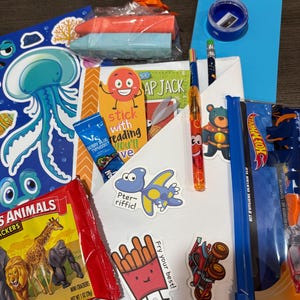 May include: A collection of children's items, including a book, crackers, stickers, pens, and a blue card. The image features a variety of colorful products, such as a blue jellyfish-themed card and a bag of mini crackers. The items are arranged on a white surface.
