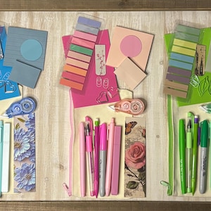 May include: A collection of stationery items in blue, pink, and green hues, arranged on a light wood-grain surface. The items include pens, sticky notes, bookmarks, and correction tape, creating a visually organized display.