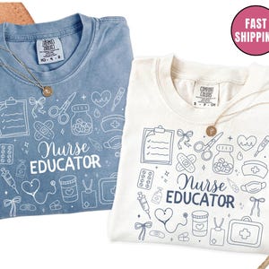 May include: Two t-shirts, one light blue and one off-white, each with a gold necklace. The shirts feature a white graphic design with medical-themed illustrations and the words "Nurse Educator". The shirts are folded.