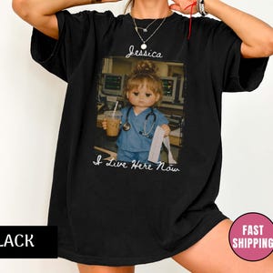 Könnte beinhalten: Ein schwarzes T-Shirt mit kurzen Ärmeln und einem Grafikdruck einer Puppe in blauer medizinischer Kleidung mit Stethoskop, die einen Kaffeebecher und einen Papierstreifen hält. Der Text "Jessica" und "I Live Here Now" ist abgebildet.
