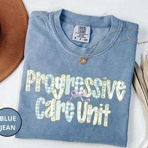 May include: A blue jean Comfort Colors t-shirt with the words "Progressive Care Unit" in a decorative font. The text features a gingham pattern and floral accents. A gold necklace and a brown hat are also visible.