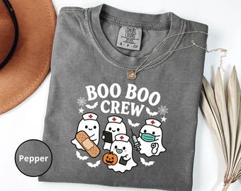 May include: Pepper-coloured long-sleeve t-shirt with the text "BOO BOO CREW" and illustrations of ghost characters dressed as nurses, holding a bandage, a tablet, a syringe, and a pumpkin. The shirt has a crew neck and a Comfort Colors tag.