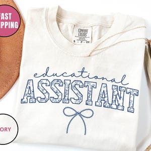 May include: Cream-coloured t-shirt featuring the text "educational ASSISTANT" in navy blue floral lettering. The shirt has a crew neck and short sleeves. A gold necklace is draped across the shirt.