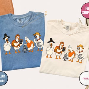 Könnte beinhalten: Zwei T-Shirts in Jeansblau und Elfenbein, die jeweils einen Grafikdruck von vier Gänsen in verschiedenen Kostümen zeigen, darunter ein Pilger, ein Truthahn, ein Koch und ein Entdecker, die mit einer kleinen grauen Roboterfigur interagieren.