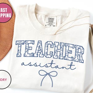 以下が含まれることがあります： アイボリー色のTシャツ。青い花柄で「TEACHER assistant」の文字がプリントされています。テキストの下には青いリボンのグラフィックがあります。シャツはクルーネックで半袖です。金のネックレスがシャツの上にかけられています。