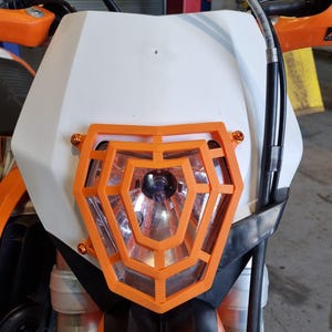 May include: Orange and black motorcycle headlight guard with a geometric design. The guard is made of plastic and is designed to protect the headlight from damage.