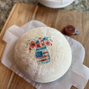 May include: A round loaf of bread dusted with flour, resting on parchment paper atop a wooden cutting board. A sticker of a mason jar with an American flag design and floral arrangement is on the bread. A white bowl and a bread lame are in the background.