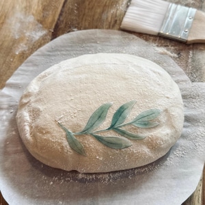May include: A round loaf of bread with a green leaf design on top, resting on a white parchment paper on a wooden surface.