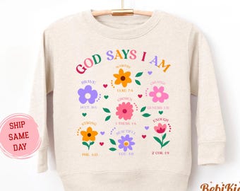 May include: A natural-colored sweatshirt with the text "GOD SAYS I AM" in colorful letters. The design includes floral graphics and inspirational words like "Brave" and "Chosen". The shirt is hanging on a wooden hanger.