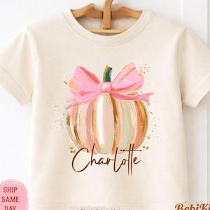 May include: A natural-colored short-sleeve t-shirt featuring a graphic of a pumpkin with a large pink bow on top. The pumpkin has gold-toned accents and glitter-like details. The name Charlotte is printed in brown script below the pumpkin.