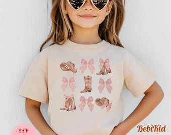 Puede incluir: Una camiseta de color natural con un diseño de acuarela de sombreros de vaquero, botas y lazos rosas. La camiseta tiene mangas cortas y la lleva un niño con gafas de sol en forma de corazón. El texto "BebiKid" es visible.