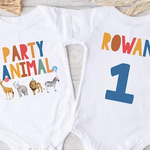 Pu&ograve; includere: Body per neonati bianco con le parole "PARTY ANIMAL" in lettere colorate e illustrazioni di animali. Il retro del body ha il nome "ROWAN" e il numero "1" in blu.