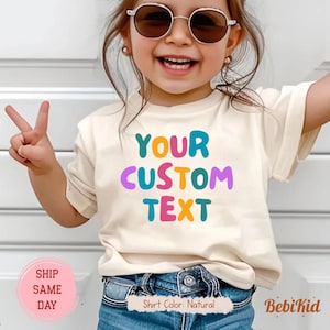 Könnte beinhalten: Ein Kind trägt ein naturfarbenes T-Shirt mit dem Text "YOUR CUSTOM TEXT" in bunten Farben. Das Kind trägt eine Sonnenbrille und eine blaue Jeans. Das T-Shirt ist von BebiKid.