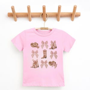 以下が含まれることがあります： カウボーイハット、リボン、ブーツの水彩デザインが施されたピンクのTシャツ。グラフィックには、茶色のカウボーイハットとブーツ、薄いピンクのリボンが含まれています。シャツは木製のハンガーに掛けられています。
