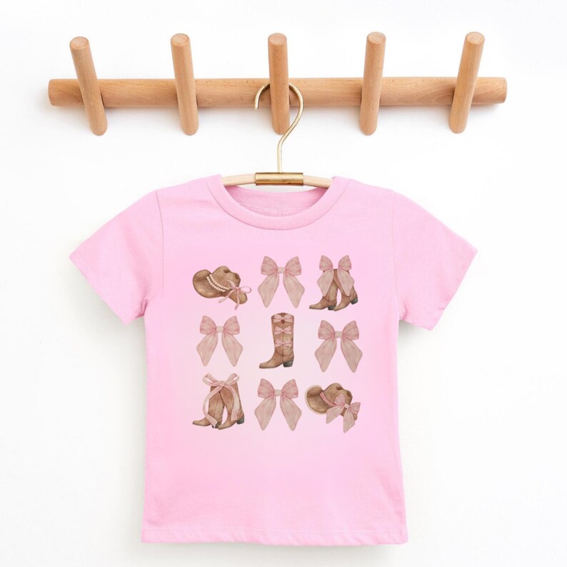 以下が含まれることがあります： カウボーイハット、リボン、ブーツの水彩デザインが施されたピンクのTシャツ。グラフィックには、茶色のカウボーイハットとブーツ、薄いピンクのリボンが含まれています。シャツは木製のハンガーに掛けられています。