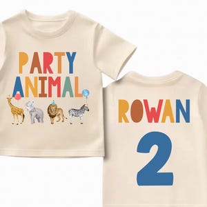 Pu&ograve; includere: Una maglietta color crema con le parole "PARTY ANIMAL" in lettere colorate e illustrazioni di animali. Il retro della maglietta presenta il nome "ROWAN" e il numero "2" in colori vivaci. Una divertente maglietta di compleanno.