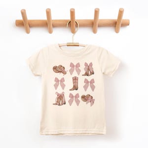 以下が含まれることがあります： クリーム色のTシャツ。茶色とピンクの色合いのカウボーイハット、リボン、ブーツの繰り返し模様が特徴です。木製のハンガーにかかっています。