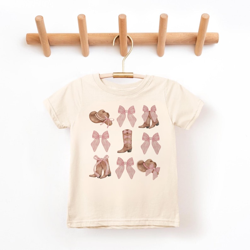 以下が含まれることがあります： クリーム色のTシャツ。茶色とピンクの色合いのカウボーイハット、リボン、ブーツの繰り返し模様が特徴です。木製のハンガーにかかっています。