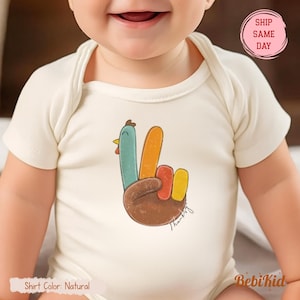 May include: A cream-coloured baby bodysuit with a colourful turkey-themed hand gesture graphic. The design includes a blue, orange, red, and yellow turkey hand with the word "Thanks" in script. The bodysuit has short sleeves and a round neckline.