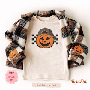 May include: A natural-colored t-shirt featuring a cartoon pumpkin wearing a gray baseball cap. The pumpkin is orange with a smiling face and is set against a black and white checkered background. A brown and cream plaid jacket is draped over the shirt.