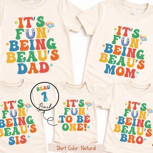 May include: A set of natural-coloured t-shirts and a onesie featuring the text "IT'S FUN BEING BEAU'S DAD", "IT'S FUN BEING BEAU'S MOM", "IT'S FUN BEING BEAU'S SIS", "IT'S FUN TO BE ONE!", and "IT'S FUN BEING BEAU'S BRO" in colourful bubble letters. One shirt's back says "BEAU 1".