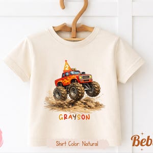 Pode incluir: Uma camiseta de cor natural com uma ilustração vibrante de um caminhão monstro vermelho com pneus grandes, um chapéu de aniversário e o número 2. O nome "Grayson" é impresso abaixo.