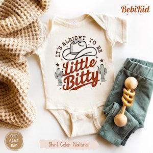 May include: Natural-colored baby onesie with the text "It's Alright to be Little Bitty" and a cowboy hat graphic. The onesie is paired with teal pants and a wooden rattle. The shirt color is natural.