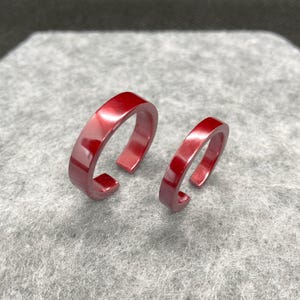 May include: Two open-ended rings with a smooth, glossy, deep red finish. The rings feature a simple, flat-band design with a gap at the back, showcasing a reflective surface that catches the light, presented in two different sizes side by side.