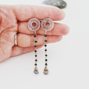 Könnte beinhalten: Silberne Ohrringe mit einem runden Design und schwarzen Perlen, die von unten hängen. Die Ohrringe sind mit einem hellblauen Edelstein verziert.