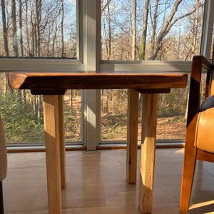 May include: A small, handcrafted wooden table with a dark brown tabletop and four light-colored wooden legs. The table is positioned in front of a large window with a view of trees. The table's dimensions are approximately 24 inches long, 18 inches wide, and 18 inches high.