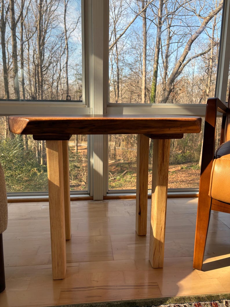 May include: A small, handcrafted wooden table with a dark brown tabletop and four light-colored wooden legs. The table is positioned in front of a large window with a view of trees. The table's dimensions are approximately 24 inches long, 18 inches wide, and 18 inches high.
