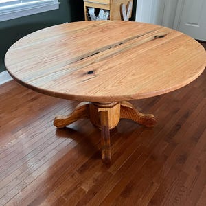 May include: A round, light brown wooden dining table with a pedestal base. The tabletop has a natural wood grain and a visible seam. The base features a central column and four curved feet. The table is approximately 48 inches in diameter.