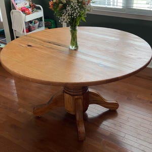 May include: A round wooden dining table with a pedestal base. The table is made of light-colored wood and has a smooth surface. A vase of flowers sits on the table. The table is approximately 48 inches in diameter.